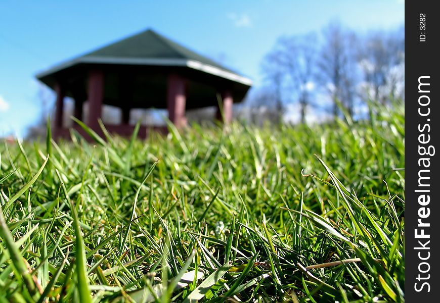 Macro of green grass, bright blue sky, trees, and gazebo in the distance. Macro of green grass, bright blue sky, trees, and gazebo in the distance.