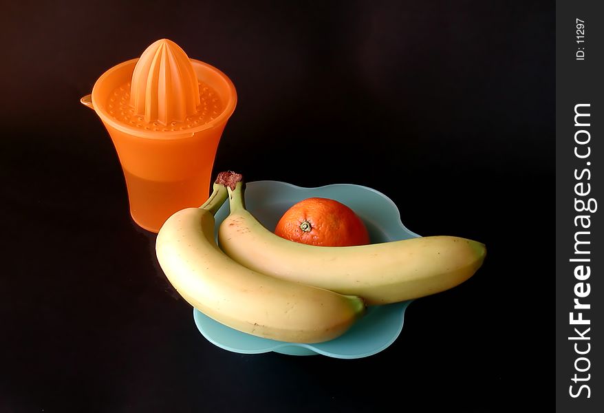 A still life composition with two fresh bananas on a blu dish and one orange, with a squeezer cup, on a black background. A still life composition with two fresh bananas on a blu dish and one orange, with a squeezer cup, on a black background