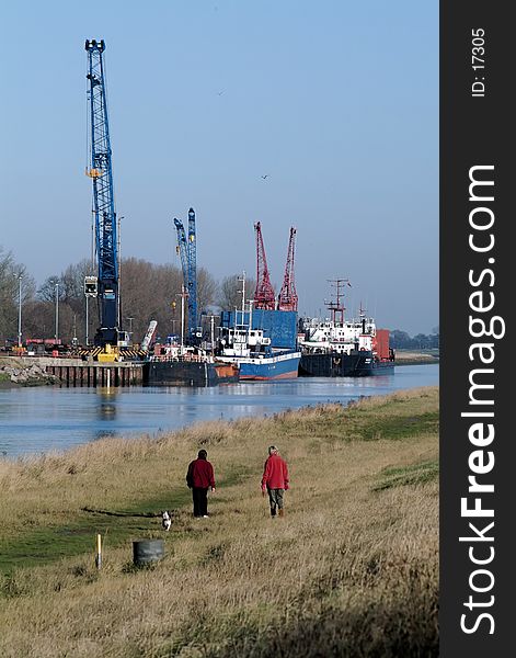 Two people walking a dog along a riverside path with a quay, ships, cranes in the background, winter, with clear blue sky backdrop. Taken at Sutton Bridge, Lincolnshire, England, UK. Two people walking a dog along a riverside path with a quay, ships, cranes in the background, winter, with clear blue sky backdrop. Taken at Sutton Bridge, Lincolnshire, England, UK