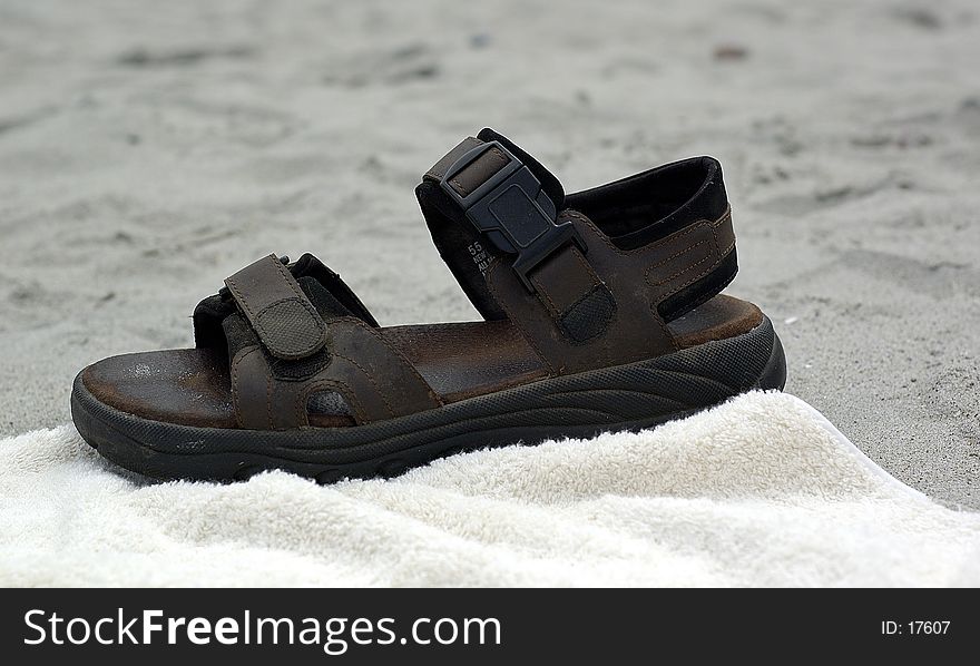 A brown sandal, sitting on a beach towel, with the grey sand behind it.