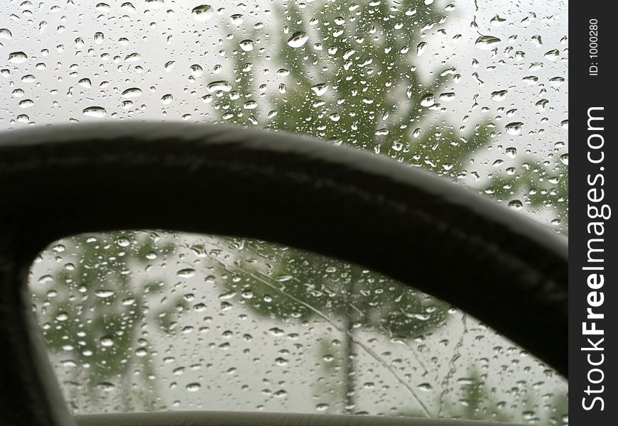 Focus on raindrops on a car windshield with steering wheel in the foreground and green trees in the background. Focus on raindrops on a car windshield with steering wheel in the foreground and green trees in the background.