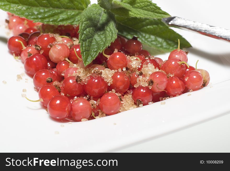 Bright red translucent edible Redcurrant berries with cane sugar and lemon balm on white plate at white background with spoon in background.