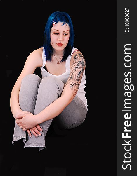 A very sad looking young girl with blue hair and a big tattoo on her lefty arm, sitting on the floor for black background, and the arms grossed around the legs. A very sad looking young girl with blue hair and a big tattoo on her lefty arm, sitting on the floor for black background, and the arms grossed around the legs.