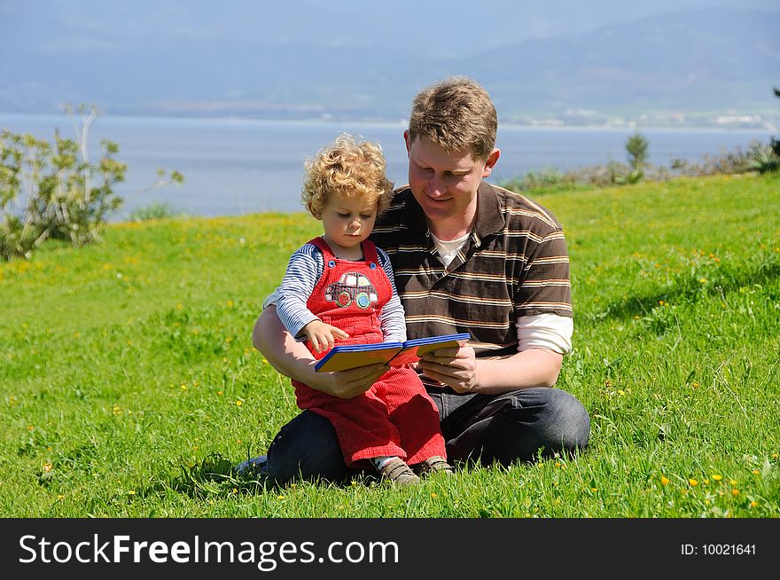 Father and son reading a book sitting on a green grass