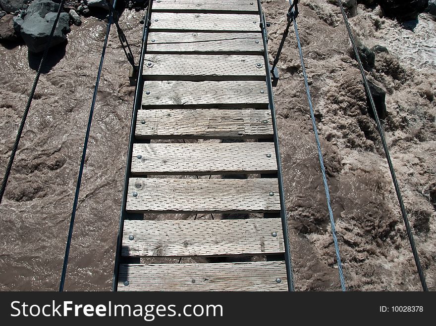 A swinging bridge crosses raging water from a glacier. A swinging bridge crosses raging water from a glacier