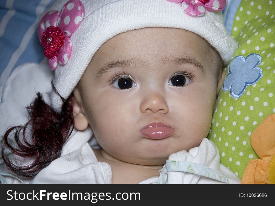 Close up face of baby who looking at camera wearing white cap with pink flower and fake hair