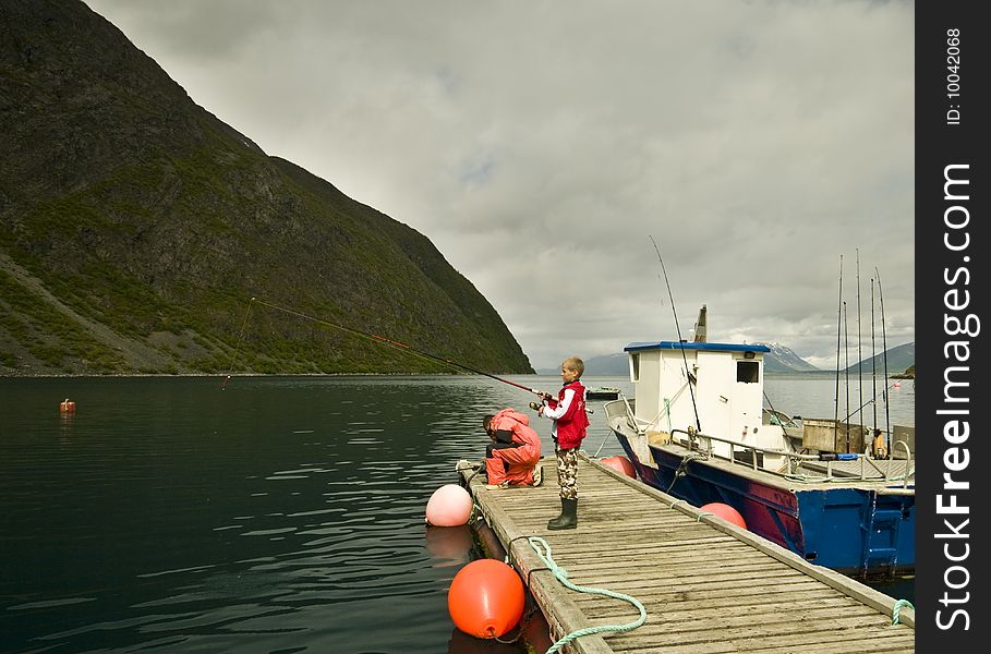 Two boys fishing in a fjord, standing on a wooden jetty next to a boat.