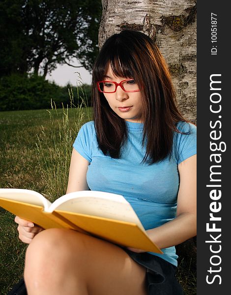 An asian girl wearing glasses sitting under a tree reading her book