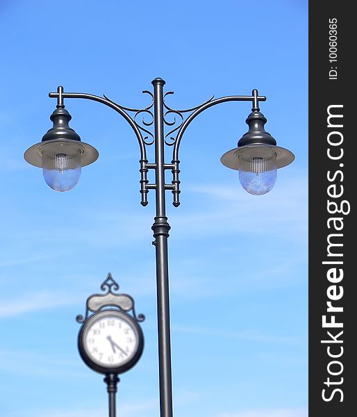 Closeup of the retro lamppost on the blue sky with blurred classic urban clock in the background. Closeup of the retro lamppost on the blue sky with blurred classic urban clock in the background.