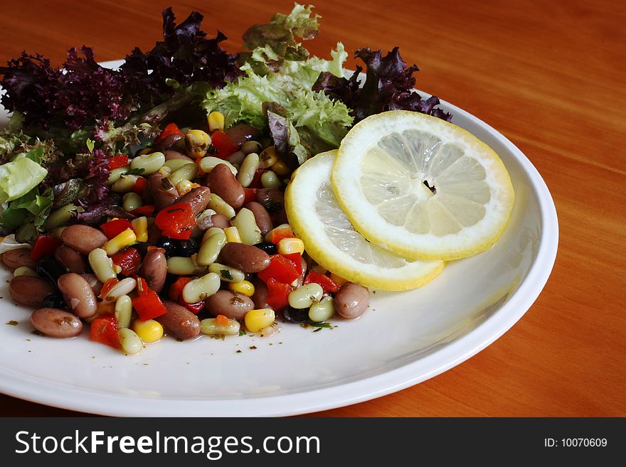 A white plate of mixed bean and lettuce salad with slices of lemon on a wood table