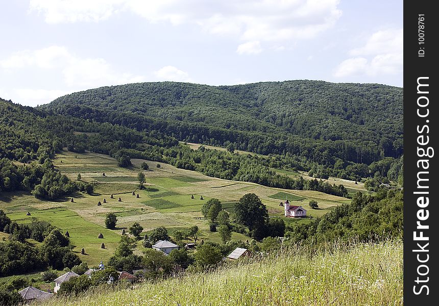 On the image there is mountain landscape,village ,field and Catholic church .