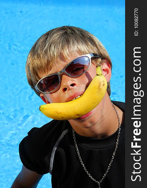 A 10 year old American - German boy with sun glasses sitting at a swimming pool in the summer sun joking with a banana between his teeth. A 10 year old American - German boy with sun glasses sitting at a swimming pool in the summer sun joking with a banana between his teeth