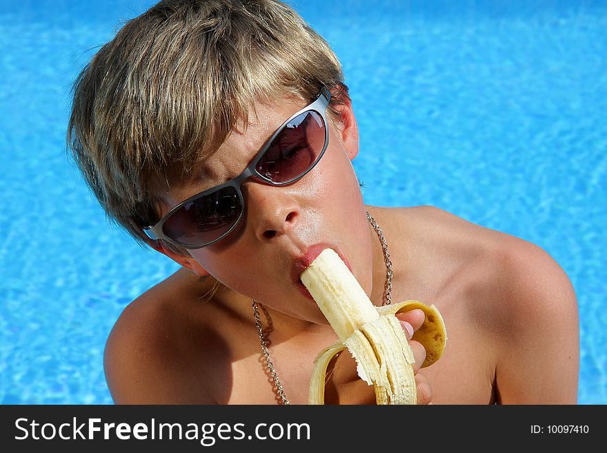 A 10 year old American - German boy with sun glasses sitting at a swimming pool in the summer sun eating a banana. A 10 year old American - German boy with sun glasses sitting at a swimming pool in the summer sun eating a banana