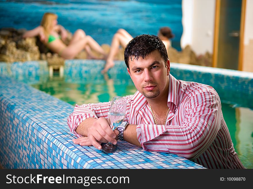 Portrait of a young man with a glass in the pool against the backdrop of two beautiful girls. Portrait of a young man with a glass in the pool against the backdrop of two beautiful girls