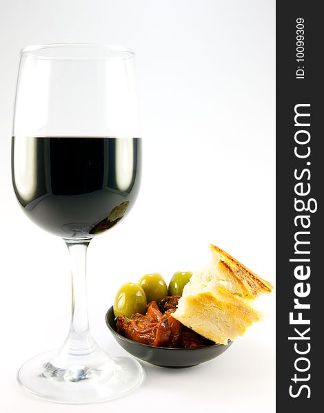 Pile of red sun dried tomatoes with three green olives and crusty bread in a small black dish with a glass of red wine on a plain background. Pile of red sun dried tomatoes with three green olives and crusty bread in a small black dish with a glass of red wine on a plain background
