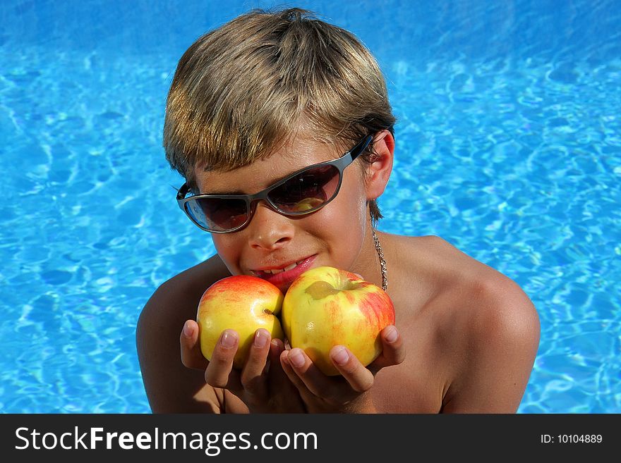 A 10 year old smiling American - German boy with sun glasses sitting at a swimming pool and presenting delicious apples in the summer sun . A 10 year old smiling American - German boy with sun glasses sitting at a swimming pool and presenting delicious apples in the summer sun