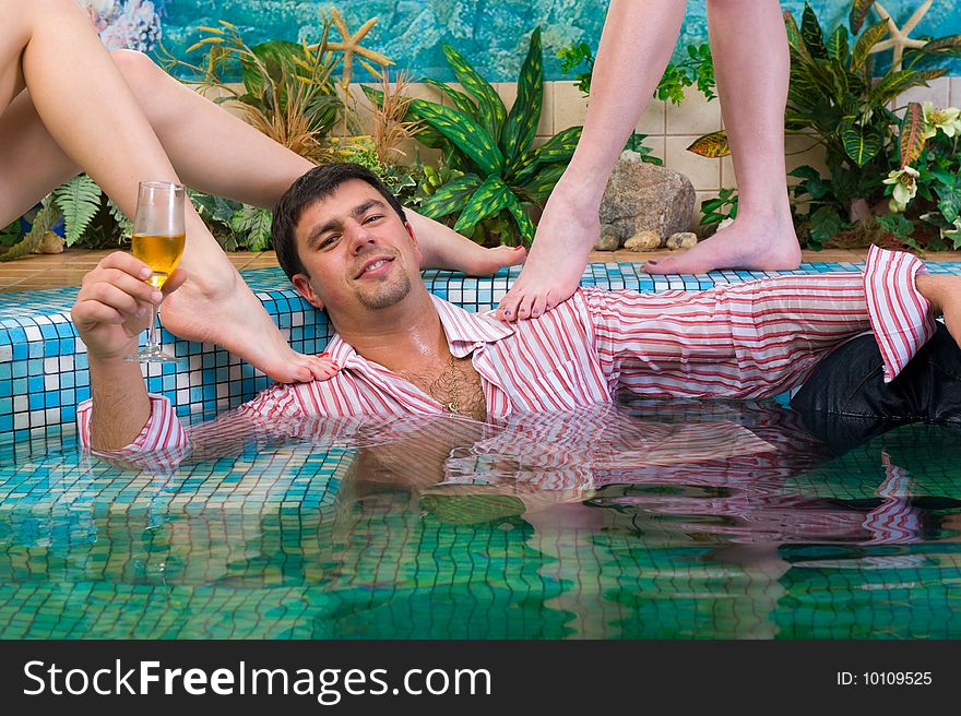 Young handsome man with a glass and the clothes in the pool surrounded by women's feet. Young handsome man with a glass and the clothes in the pool surrounded by women's feet
