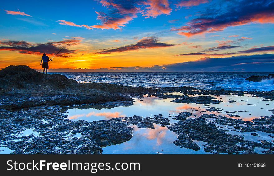 Dramatic orange and pink sunset with lone fisherman seen in silhouette using rod and line fishing from rocks on the seashore of Taiwan. Dramatic orange and pink sunset with lone fisherman seen in silhouette using rod and line fishing from rocks on the seashore of Taiwan.