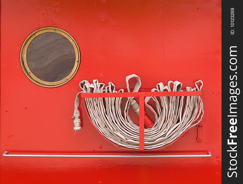 Closely cropped details on a red metal wall of a tugboat showing fire hose and round window. Closely cropped details on a red metal wall of a tugboat showing fire hose and round window