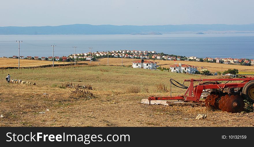 White and yellow homes at the coast of the blue sea with islands, in a pastoral setting behind harvested fields, sheep and shepherd, disc-harrow farm equipment. Electric poles lined through the road between fields. Gulf of Saros, northern part of the Egean Sea. White and yellow homes at the coast of the blue sea with islands, in a pastoral setting behind harvested fields, sheep and shepherd, disc-harrow farm equipment. Electric poles lined through the road between fields. Gulf of Saros, northern part of the Egean Sea.