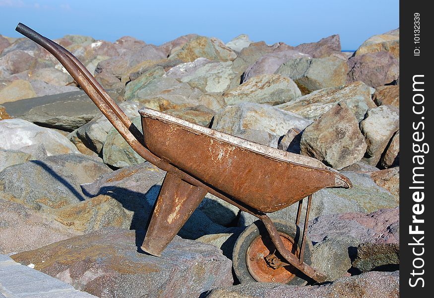Old and rusted wheel barrow with one wheel placed above large blocks of rocks under blue sky. Old and rusted wheel barrow with one wheel placed above large blocks of rocks under blue sky