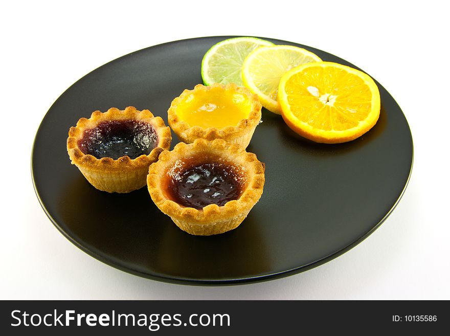 Red and yellow small jam tarts with slices of lemon, lime, and orange on a black plate on a white background. Red and yellow small jam tarts with slices of lemon, lime, and orange on a black plate on a white background