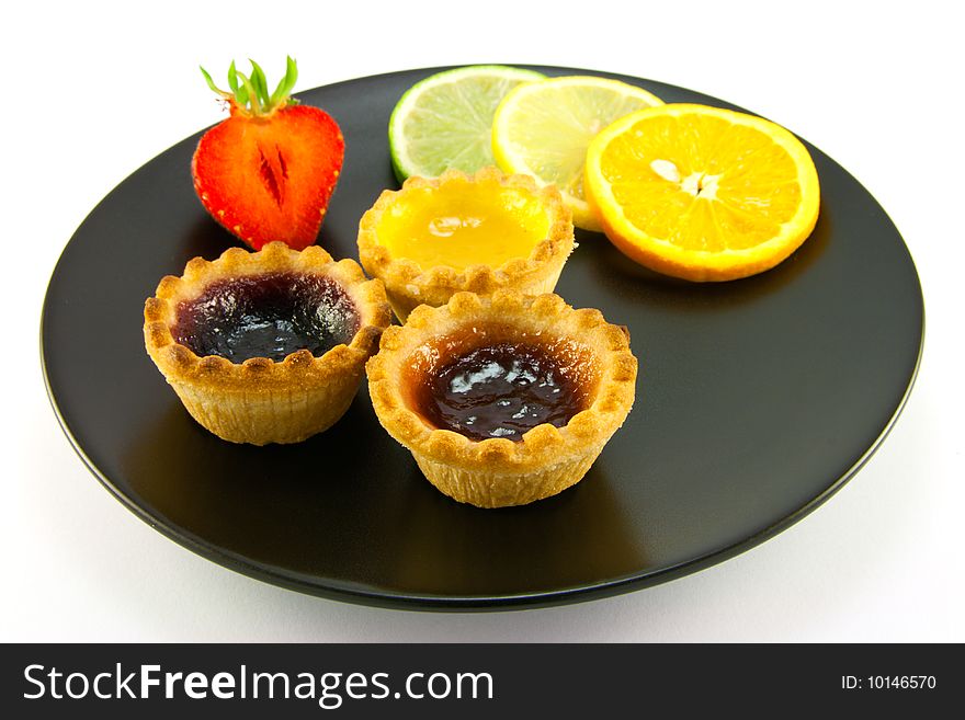 Red and yellow small jam tarts with slices of lemon, lime, orange and strawberry on a black plate on a white background. Red and yellow small jam tarts with slices of lemon, lime, orange and strawberry on a black plate on a white background