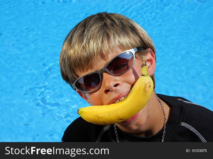 A 10 year old American - German boy with sun glasses joking with a banana in his mouth sitting at a swimming pool in the summer sun. A 10 year old American - German boy with sun glasses joking with a banana in his mouth sitting at a swimming pool in the summer sun