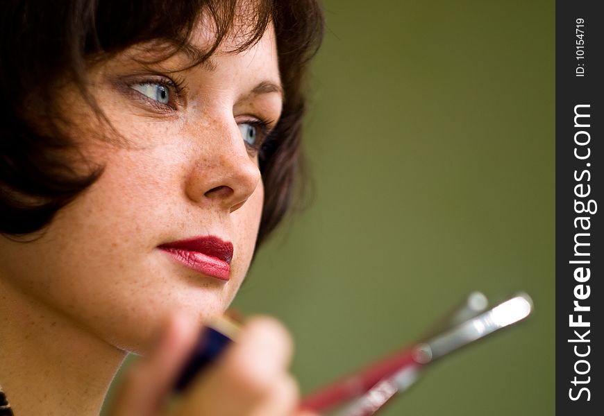 Brown-haired girl makes a make-up. Looks aside. Green background. Lipstick red. Eyes blue. Holds pencils. Low depth of field. Brown-haired girl makes a make-up. Looks aside. Green background. Lipstick red. Eyes blue. Holds pencils. Low depth of field.