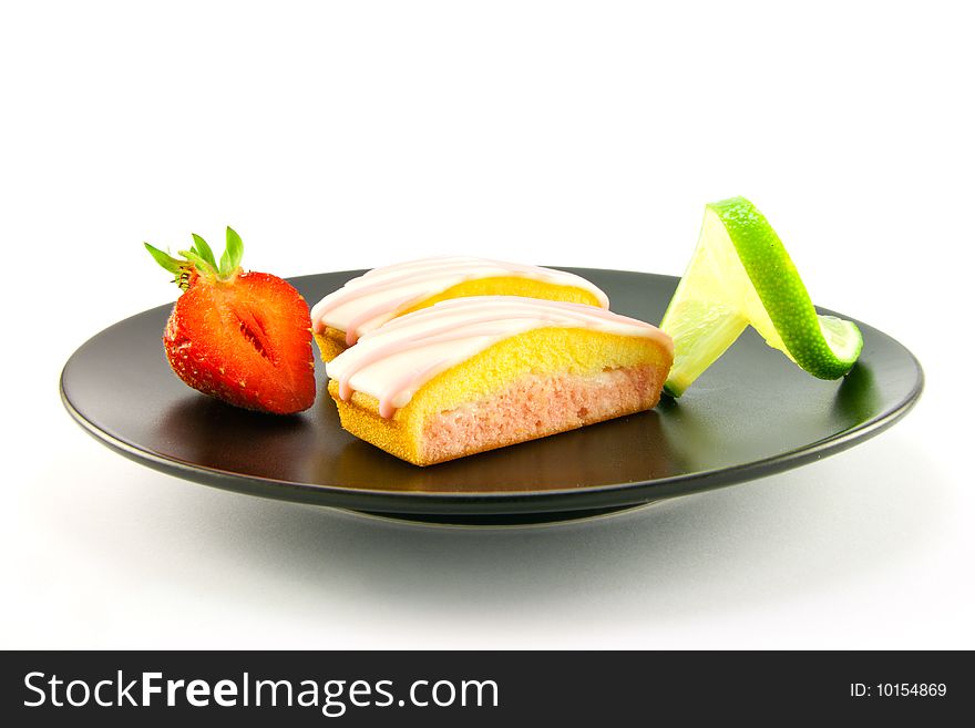 Two pink strawberry sliced cakes with icing on the top with a half strawberry and twist of lime on a black plate on a white background. Two pink strawberry sliced cakes with icing on the top with a half strawberry and twist of lime on a black plate on a white background