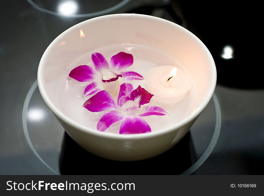 A white porcelain bowl with water, two purple orchids and a floating tealight candle on a black surface. A white porcelain bowl with water, two purple orchids and a floating tealight candle on a black surface.