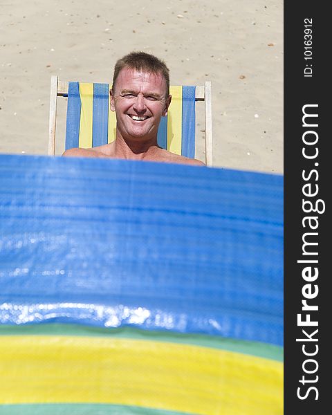 A happy smiling forties man sitting on a stripped deckchair in front of a windbreak on a sandy beach. A happy smiling forties man sitting on a stripped deckchair in front of a windbreak on a sandy beach.