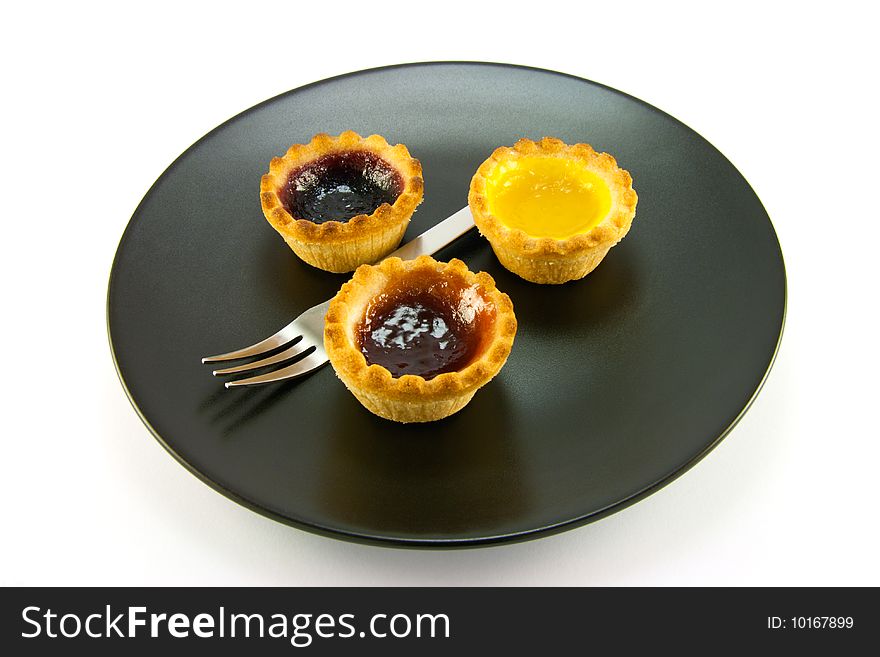 Three red and yellow small pastry jam tarts on a black plate with a fork on a white background. Three red and yellow small pastry jam tarts on a black plate with a fork on a white background