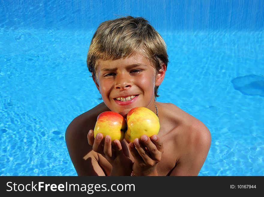 A 10 year old cute and smiling American - German boy sitting at a swimming pool and presenting delicious apples in the summer sun. A 10 year old cute and smiling American - German boy sitting at a swimming pool and presenting delicious apples in the summer sun