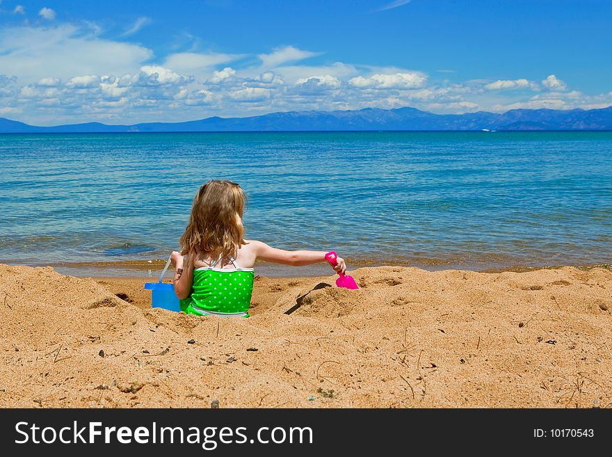 Young girl sitting in the sand facing the lake playing in the sand. Young girl sitting in the sand facing the lake playing in the sand.