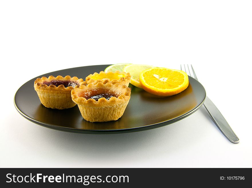 Red and yellow small jam tarts with slices of lemon, lime, and orange on a black plate with a fork on a white background. Red and yellow small jam tarts with slices of lemon, lime, and orange on a black plate with a fork on a white background