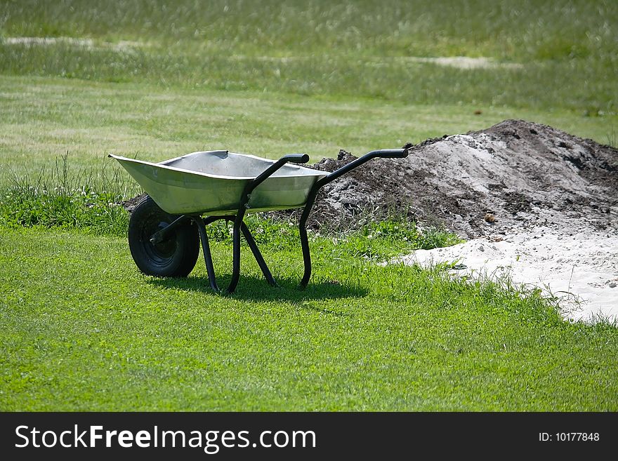 The wheelbarrow is standing on the grass field near a heap of sand. The wheelbarrow is standing on the grass field near a heap of sand