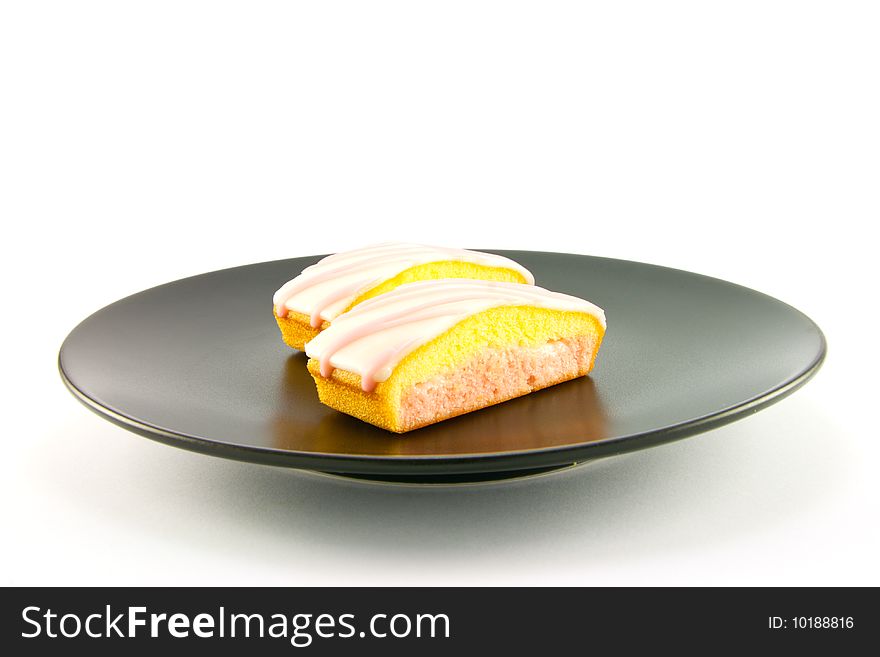 Two strawberry sliced cakes with icing on the top and pink and yellow sponge on a black plate with a white background. Two strawberry sliced cakes with icing on the top and pink and yellow sponge on a black plate with a white background