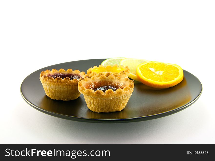 Red and yellow small jam tarts with slices of lemon, lime, and orange on a black plate on a white background. Red and yellow small jam tarts with slices of lemon, lime, and orange on a black plate on a white background