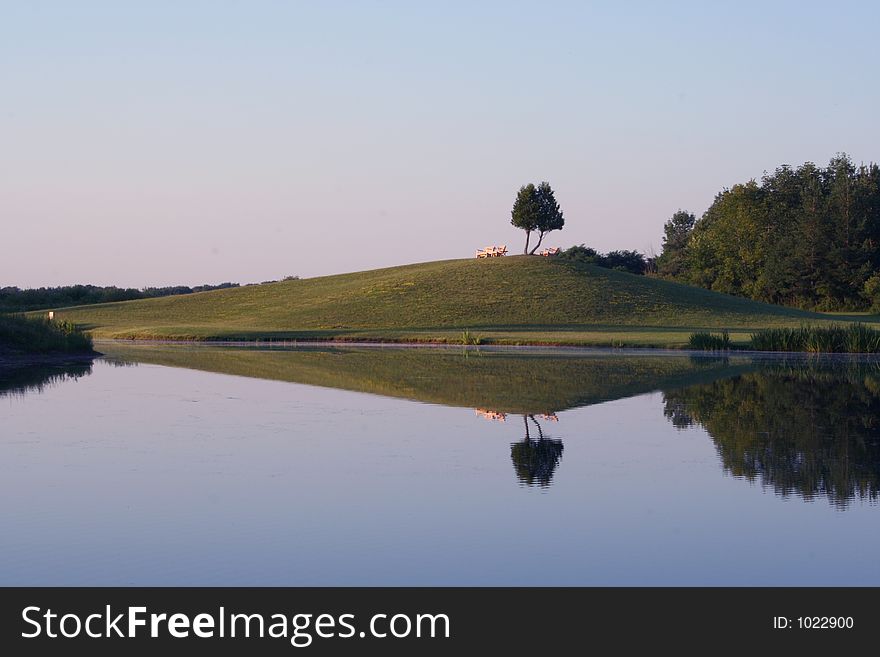 A pair of benches sit on top of a hill all reflected in a still pond at sunset. A pair of benches sit on top of a hill all reflected in a still pond at sunset