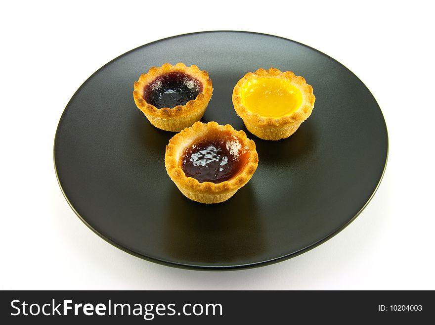 Three red and yellow small pastry jam tarts on a black plate with a white background. Three red and yellow small pastry jam tarts on a black plate with a white background