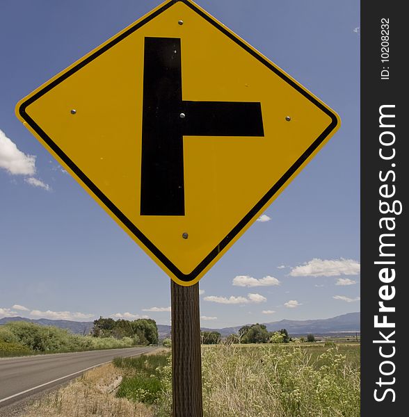 This is a road sign with a highway, mountains and a bright blue sky in the backgraound.