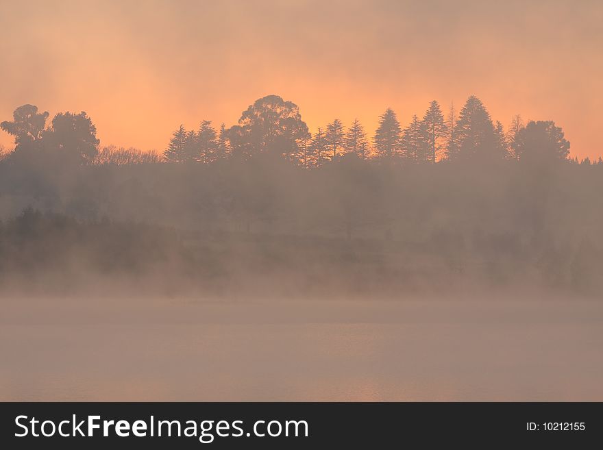 Early morning at a lake/dam with an orange glow visible through the trees and mist. Early morning at a lake/dam with an orange glow visible through the trees and mist