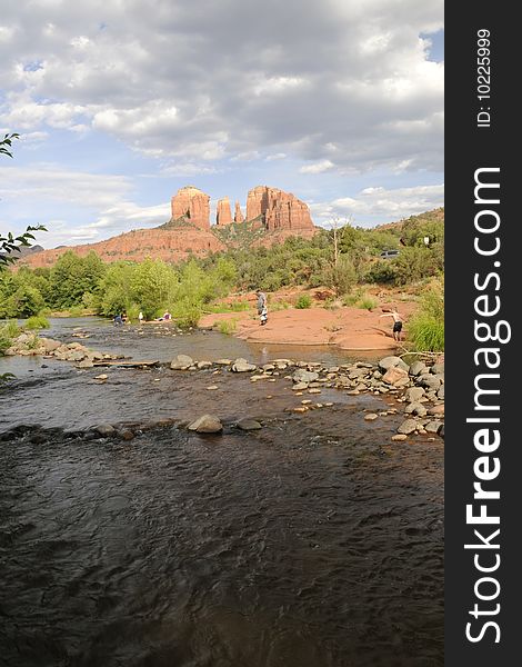 View of red rock formation, cathedral rock in sedona arizona, with small river running in the foregraound.  various people hiking and playing along riverbank