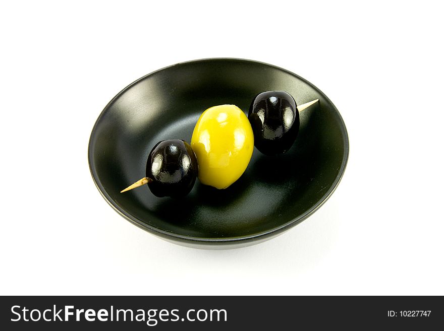 Two black and one green olive on a stick in a small black bowl on a white background. Two black and one green olive on a stick in a small black bowl on a white background