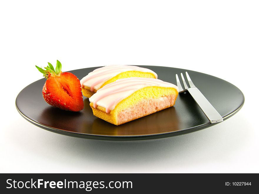 Two pink strawberry sliced cakes with icing on the top with a half strawberry and small fork on a black plate with a white background. Two pink strawberry sliced cakes with icing on the top with a half strawberry and small fork on a black plate with a white background