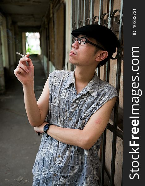 Bespectacled asian man in beret smoking while waiting along a corridor in a deserted abandoned place. Bespectacled asian man in beret smoking while waiting along a corridor in a deserted abandoned place