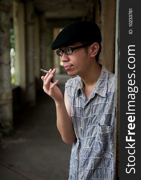 Frowning bespectacled asian man in beret smoking while waiting along a corridor in a deserted abandoned place