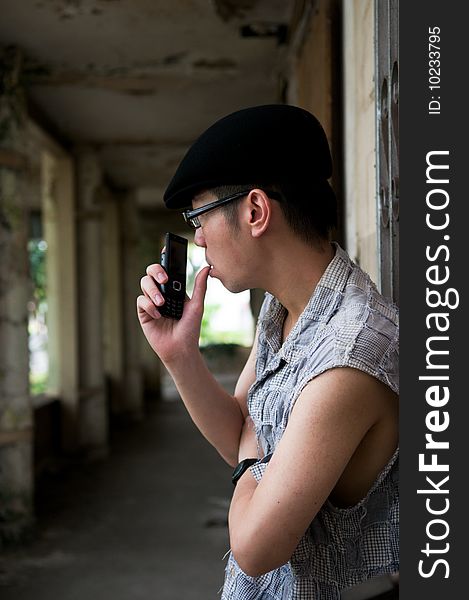 Bespectacled asian man in beret holding a mobile phone and with thumb on lips along a corridor in a deserted abandoned place. Bespectacled asian man in beret holding a mobile phone and with thumb on lips along a corridor in a deserted abandoned place