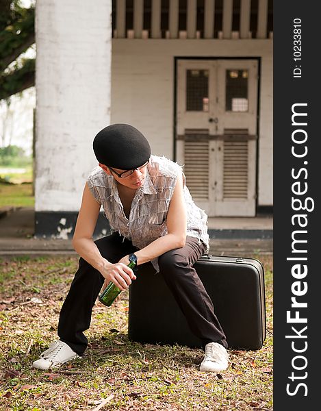 Bespectacled asian man in beret holding a bottle and sitting on a suitcase outside the boarded up door of an abandoned apartment. Bespectacled asian man in beret holding a bottle and sitting on a suitcase outside the boarded up door of an abandoned apartment